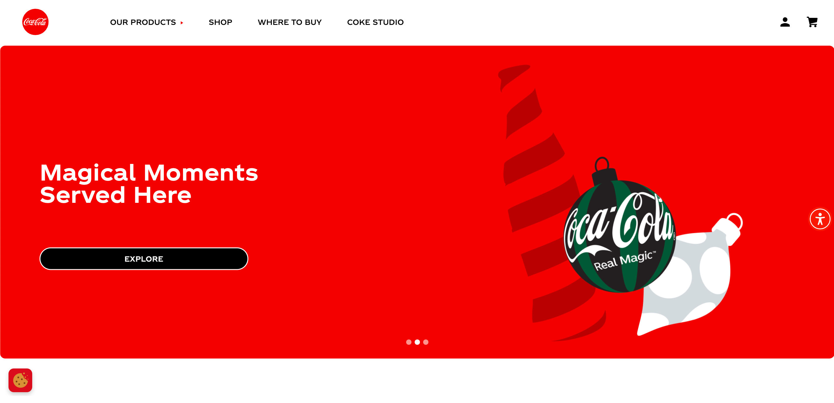 The Coca-Cola website homepage. The menu bar at the top of the page has a white background with black text. The red Coca-Cola logo is positioned on the far left of the menu bar. Below the menu is a large background with white text and a graphic of a green and black bauble.